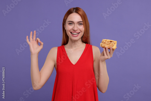 Young woman with piece of tasty cake showing ok gesture on purple background