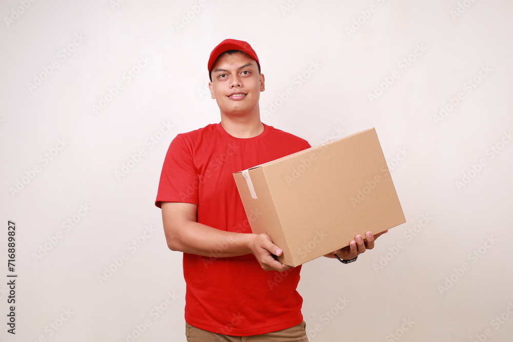 Happy Asian young courier man in red standing with cardboard box. Isolated on white