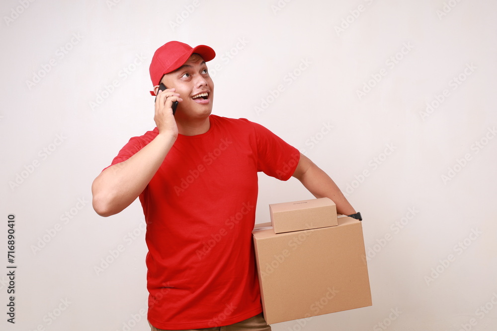 Excited Asian courier man in red holding a cardboard box while talking on the phone and looking above Excited Asian courier man in red holding a cardboard box while talking on the phone and looking above