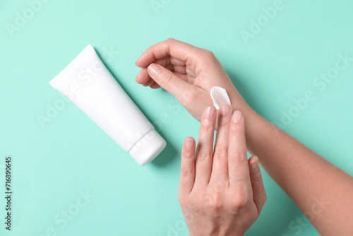 Woman with tube applying cosmetic cream onto her hand on turquoise background, top view