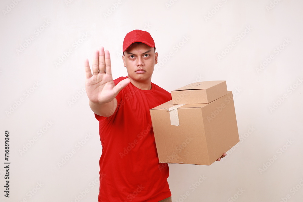 Asian courier man in red holding a cardboard box while showing rejection gesture. Isolated on white Asian courier man in red holding a cardboard box while showing rejection gesture. Isolated on white