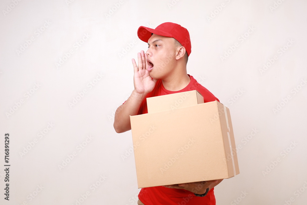 Asian courier man in red shouting to sideways while holding a cardboard box. Isolated on white Asian courier man in red shouting to sideways while holding a cardboard box. Isolated on white