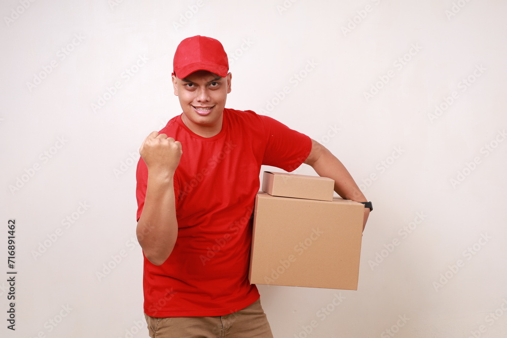 Excited Asian courier man in red celebrating success with fist hand while holding a cardboard box. Isolated on white Excited Asian courier man in red celebrating success with fist hand while holding a cardboard box. Isolated on white