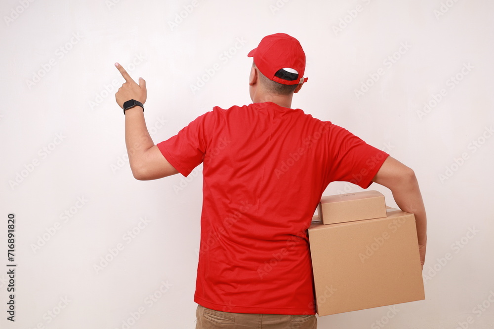Back view of Asian courier man in red pointing sideways while holding a cardboard box. Isolated on white Back view of Asian courier man in red pointing sideways while holding a cardboard box. Isolated on white