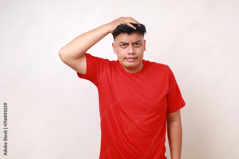 Confused asian young man in red standing while holding his head and looking at the camera Confused asian young man in red standing while holding his head and looking at the camera