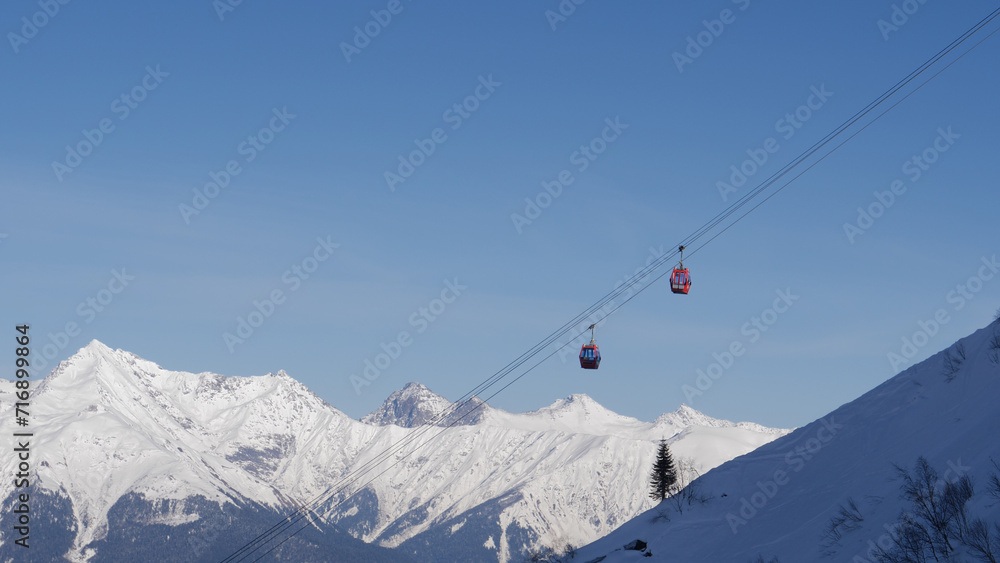 Ski resort on sunny winter day. Ski lift funicular cab lifts skiers and snowboarders up mountain against clear blue sky. Breathtaking view of snowy peaks of mountains and valleys of glaciers.