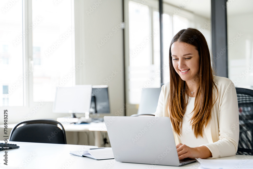 A young businesswoman is captured in a candid moment of joy while working on her laptop, sitting at her office desk. Pleasant aspects of office life and the satisfaction that comes from engaging work. A young businesswoman is captured in a candid moment of joy while working on her laptop, sitting at her office desk. Pleasant aspects of office life and the satisfaction that comes from engaging work.