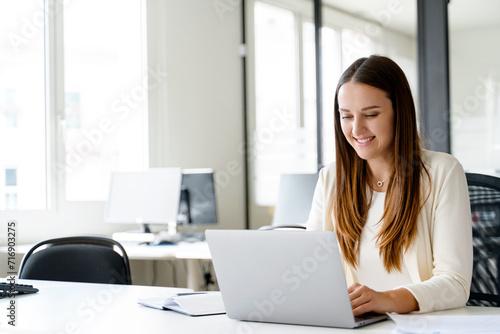 A young businesswoman is captured in a candid moment of joy while working on her laptop, sitting at her office desk. Pleasant aspects of office life and the satisfaction that comes from engaging work.