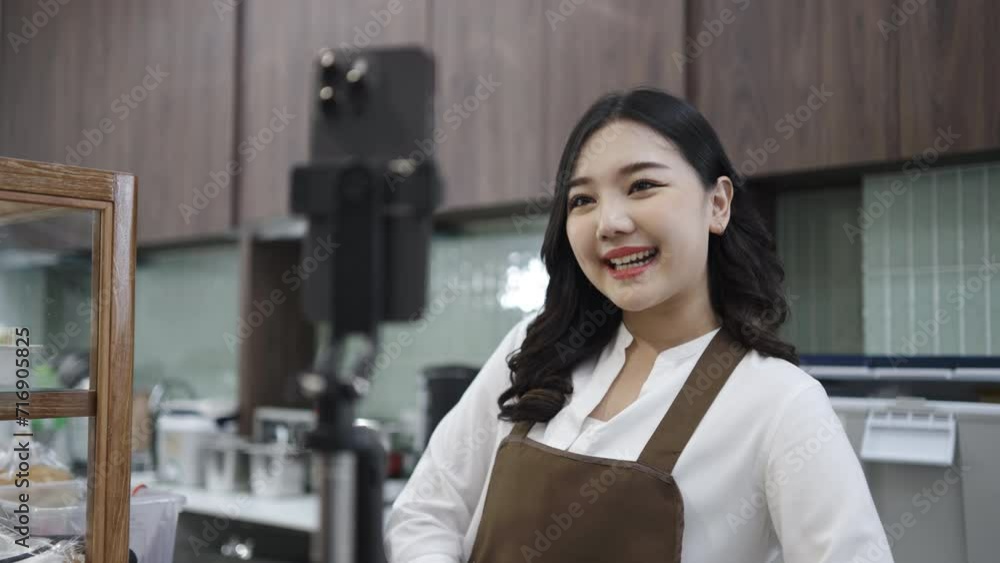 Young asian women is shop owner put on an apron show bread packages for sale while live streaming or recording video on social media in her cafe, coffee shop business concept. Young asian women is shop owner put on an apron show bread packages for sale while live streaming or recording video on social media in her cafe, coffee shop business concept.