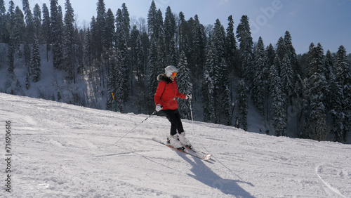 Sports woman skier in bright red sportswear skiing down snowy slope in mountains at ski resort. Mountain scenery with snow capped hills with pine forest. Winter recreation and sports. Shot in motion