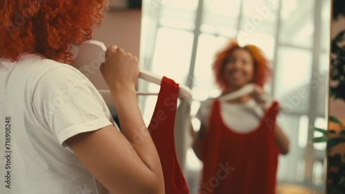 Red-haired woman getting ready for a party, choosing between dresses near mirror photo