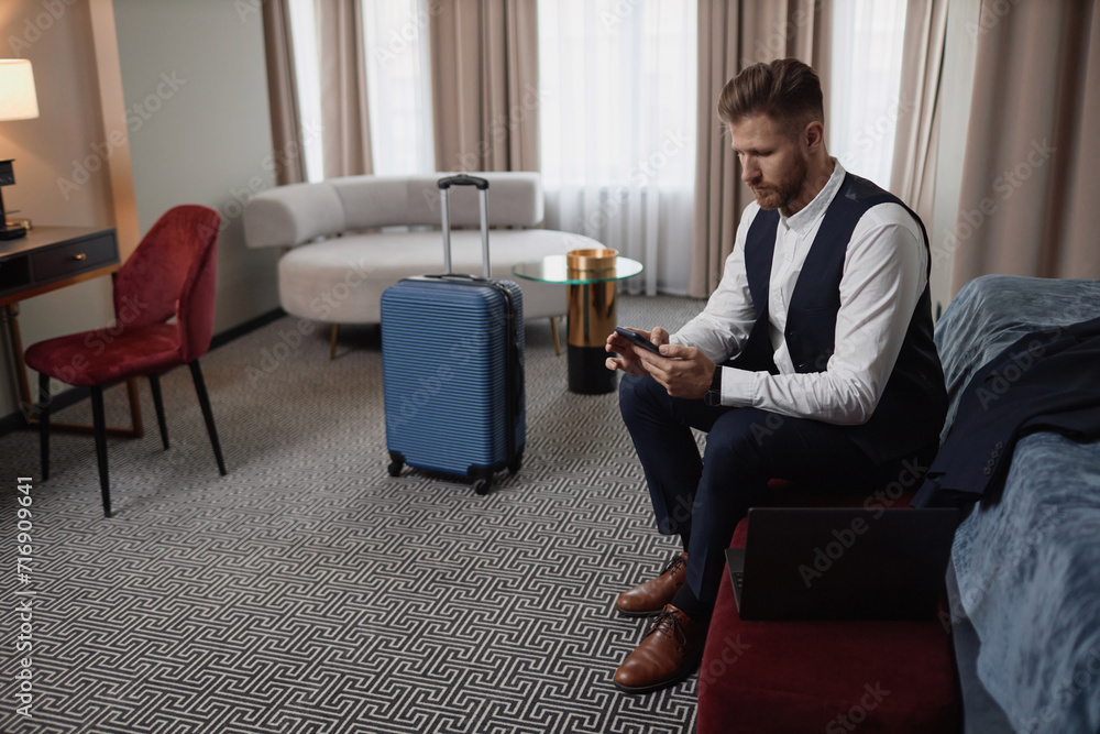 Full length portrait of elegant young businessman using smartphone in hotel room with suitcase in background, copy space Full length portrait of elegant young businessman using smartphone in hotel room with suitcase in background, copy space