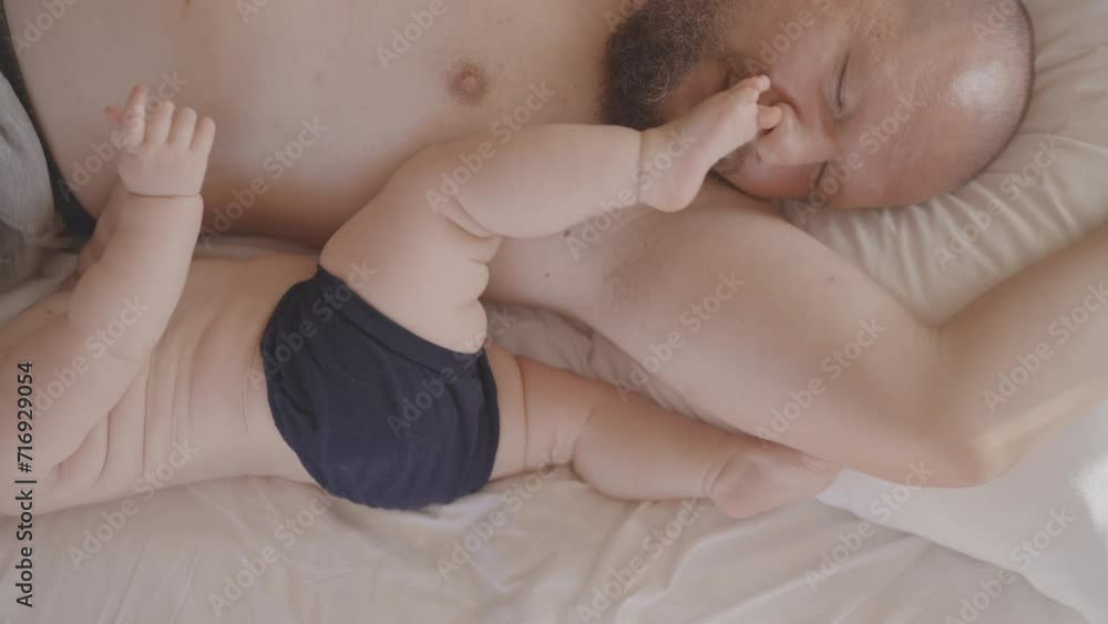 Young tired father sleep near little baby child in a bed. Child feet waking up daddy. Concept of family, parenthood, childhood. Touching tender moment of love, soft touch. Authentic shot loving dad Young tired father sleep near little baby child in a bed. Child feet waking up daddy. Concept of family, parenthood, childhood. Touching tender moment of love, soft touch. Authentic shot loving dad