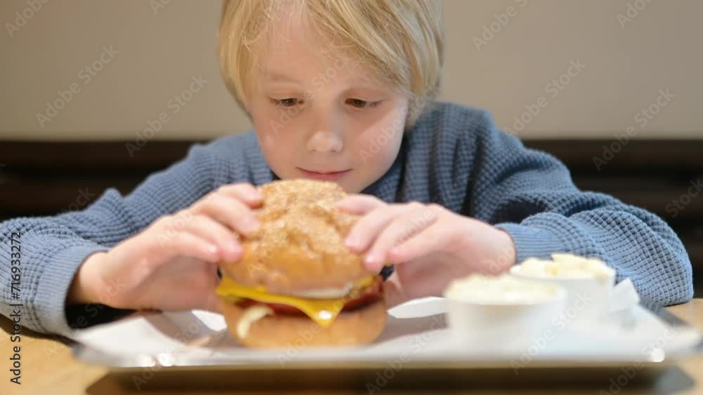 Cute blonde preteen boy eating large hamburger at fast food restaurant. Unhealthy meal for kids. Junk food. Overweight problem child. Cute blonde preteen boy eating large hamburger at fast food restaurant. Unhealthy meal for kids. Junk food. Overweight problem child.