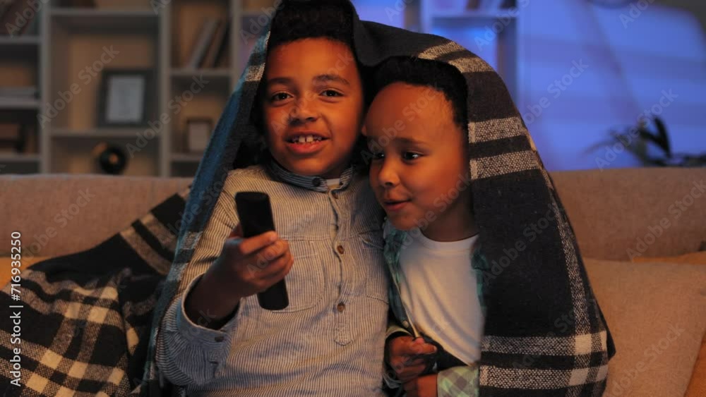 Two little African American brothers are sitting on the sofa covered with a blanket and watching TV. Two multiracial boys get scared while watching a horror movie. Two little African American brothers are sitting on the sofa covered with a blanket and watching TV. Two multiracial boys get scared while watching a horror movie.