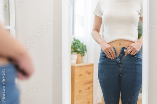Excess weight gain, overweight, close up young woman wearing fit trousers standing looking mirror at home, hand trying to close zip up and button of jeans from fat around waist. Obese problem concept. photo