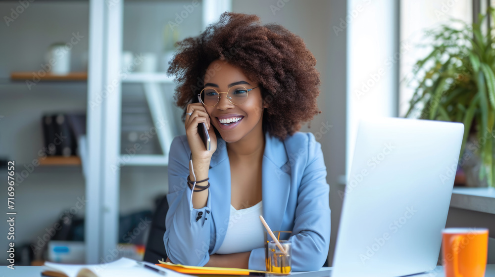 Obraz premium Smiling young woman speaking on a smartphone in a well-lit, modern office setting with a laptop