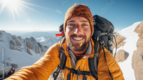 Man in orange jacket taking a selfie on a snowy mountain with clear blue skies.