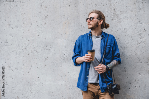 Portrait of cheerful young caucasian man holding coffee cup and looking away while standing against grey background. Modern freelancer tourist hipster walking outdoors photo