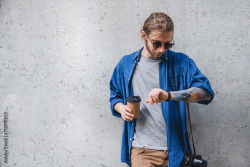 Portrait of cheerful young caucasian man holding coffee cup and checking time on watch while standing against grey background. Modern freelancer walking in the city