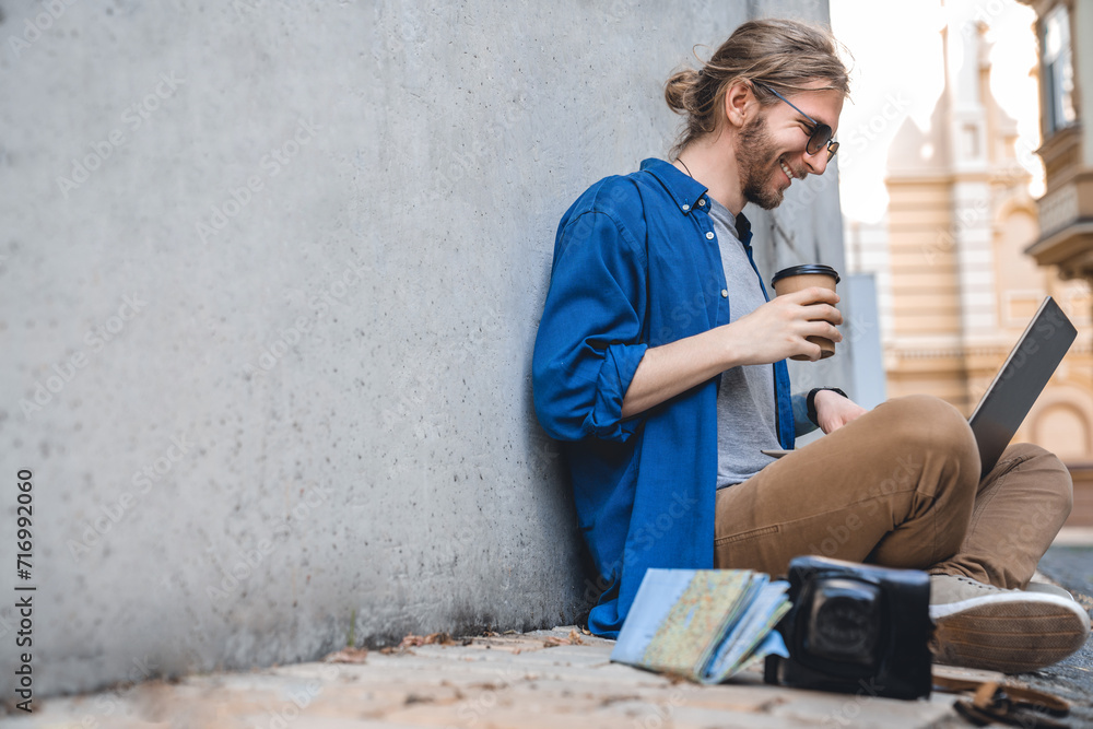 Side view shot of young stylish man work using laptop outdoors with coffee cup in hand. The concept of study, freelancing, work and travel. Modern student typing, doing projects online