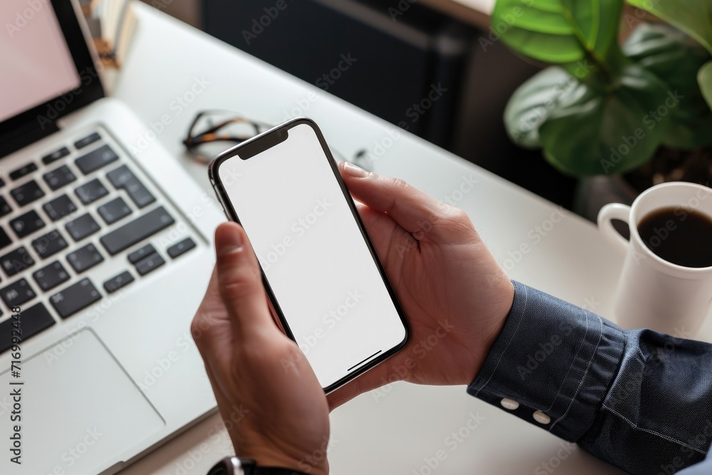 Close-up of a person hands holding a blank screen smartphone above a modern white office desk with laptop and accessories.. Close-up of a person hands holding a blank screen smartphone above a modern white office desk with laptop and accessories..