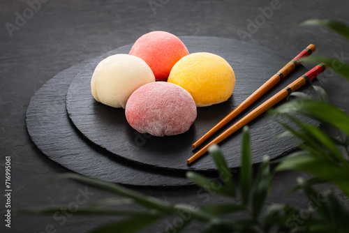 Four round multi-colored mochi lie on black slate. Dark background with leaves and sticks. Perfect retouching. Advertising. View from above. Photo