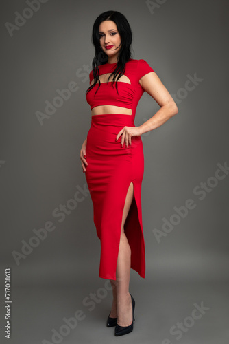 Studio portrait of young beautiful smiling woman in evening red dress with black loose hair against fashion gray background.