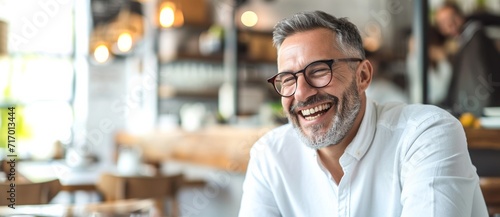 A jovial man in glasses chuckles heartily, his bearded face framed by the stylish indoor decor of a cozy restaurant photo