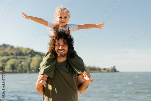 Child daughter sits on her dad shoulders outdoors on a summer beach and looking at camera