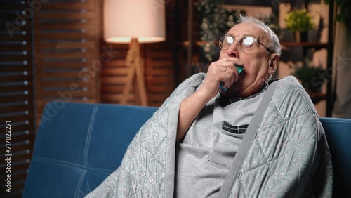 Portrait of old man with COVID or influenza virus in home, person coughing and wrapping in blanket photo