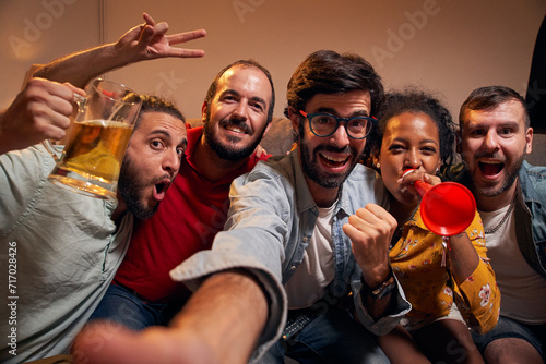 Selfie portrait looking at camera of group handsome Caucasian friendly women watching football game on TV while sitting on sofa at night. Friends looking at camera POV sport channel late in evening.