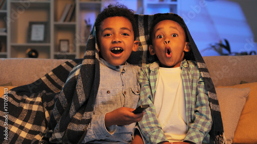 Two little African American brothers are sitting on the sofa covered with a blanket and watching TV. Two multiracial boys get scared while watching a horror movie.