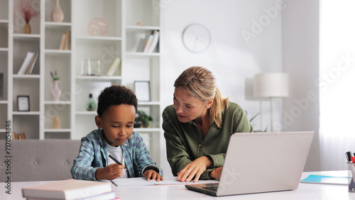 Blonde tutor teaching little African American preschooler to write at home. Help with early childhood education in an online school. Happy boy is learning to write. photo