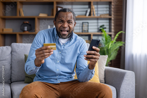African american man sitting on sofa while holding mobile phone and plastic bank card with stressed facial expression. Adult male buyer making unsuccessful attempts paying for online order at home.