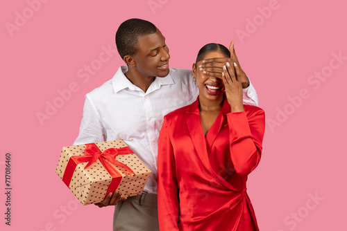 Black man surprises woman with gift, covering her eyes, both in formal attire photo