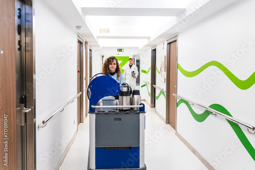 A warm-hearted nurse behind a coffee service cart in a hospital corridor, with a doctor in the soft-focused background.