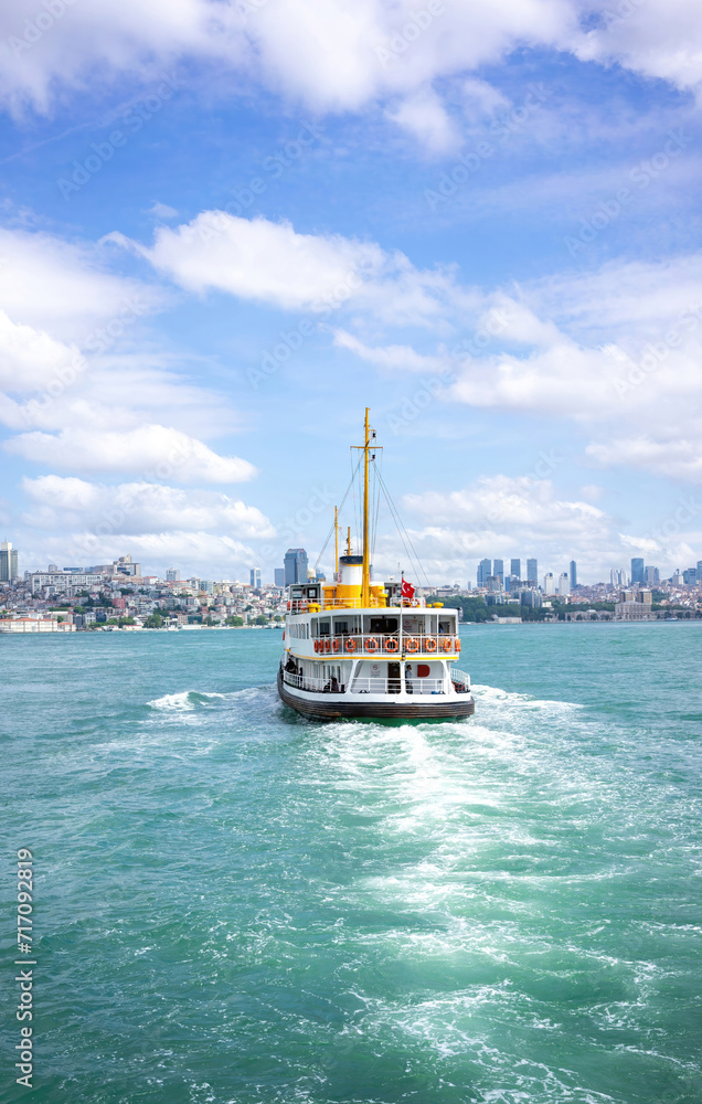 Obraz premium A classic ferry boat cuts through the turquoise waters, connecting the vibrant cityscape under a sky dotted with fluffy clouds