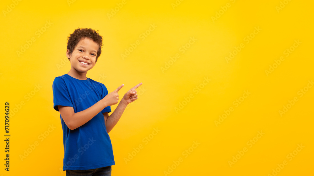 Latin boy with bright smile pointing at copy space against yellow backdrop