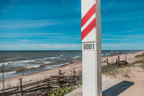 Red and white striped border post marked '0001' on a sunny beach, symbolizing the starting point of the border along the coastline
