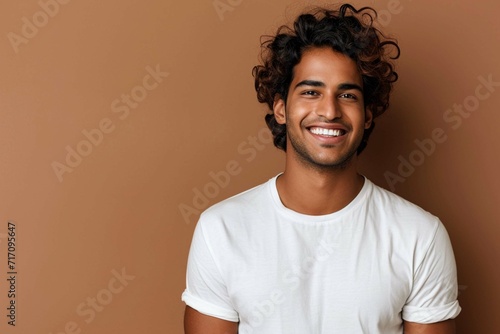 Portrait of happy Hindu man with curly hair smile toothily keeps arms down dressed in casual t shirt looks cheerful isolated over brown background being in good mood. Human positive emotions concept © Manzoor