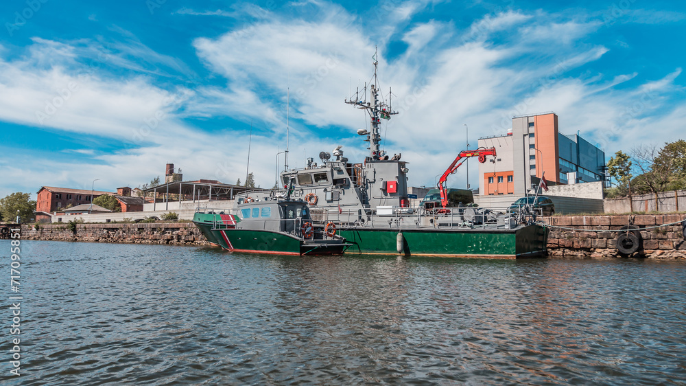 Naval patrol boat moored at the city dock with industrial equipment on board against a backdrop of urban buildings and a clear sky