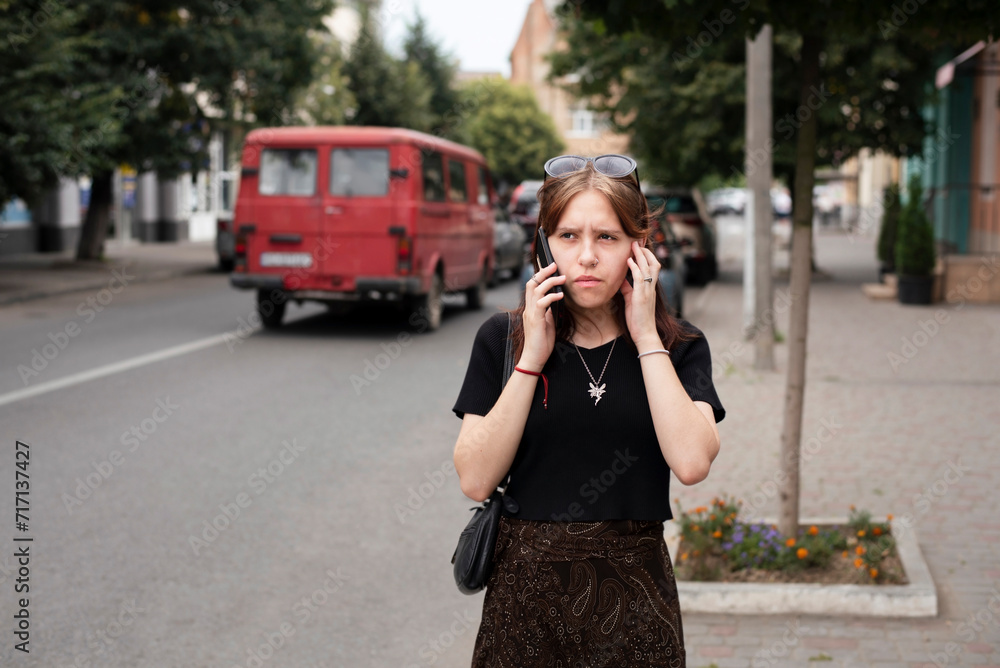 A young teenage girl is talking on the phone in the city but cannot hear her interlocutor because it is very noisy in the city, a girl is walking in city and talking on phone