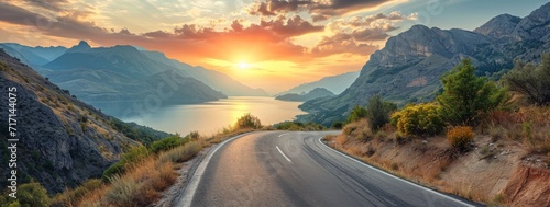 A winding road through a majestic mountain pass leads to a tranquil lake surrounded by towering trees, under a colorful sky at sunrise and sunset