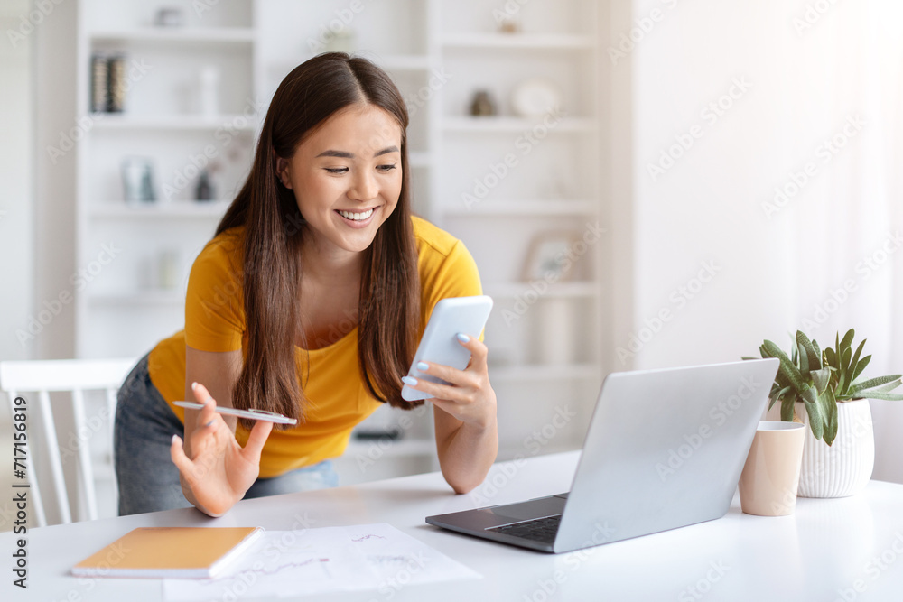 Cheerful asian woman using smartphone while working at desk in home office Cheerful asian woman using smartphone while working at desk in home office