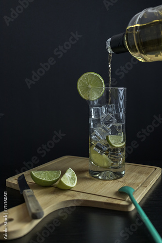 Piscola, preparing, cocktail, adding, pisco, glass with ice, wooden board, cut lemons, knife, spoon. On table and black background. from Chile, summer, vertical. Typical Chilean food and drink