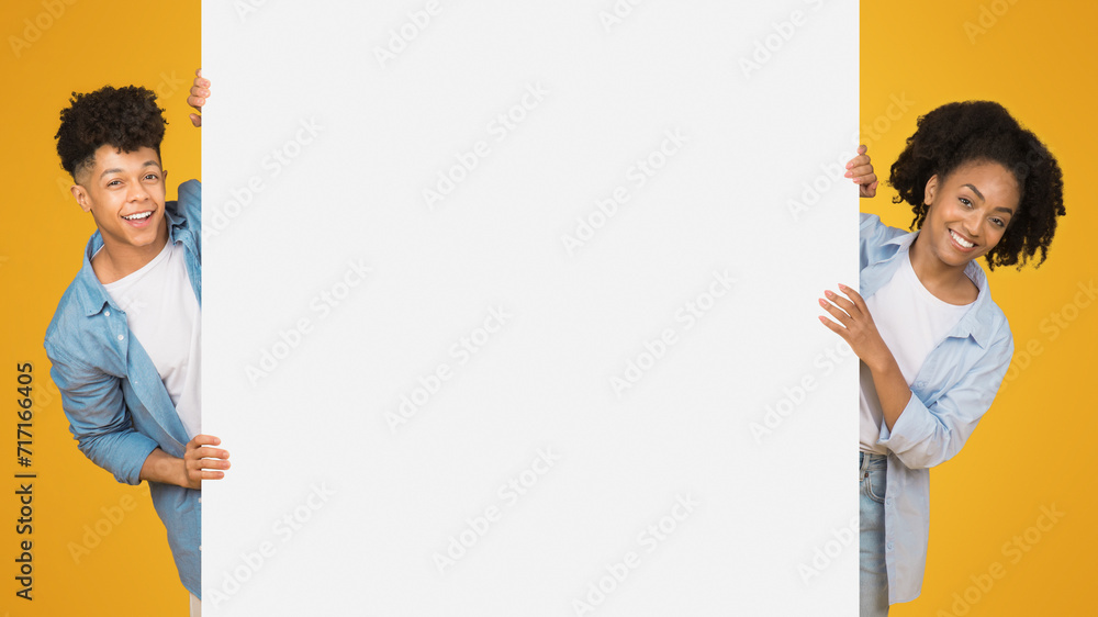 Cheerful young woman and man, peeking from behind a blank white banner with plenty of copy space Cheerful young woman and man, peeking from behind a blank white banner with plenty of copy space