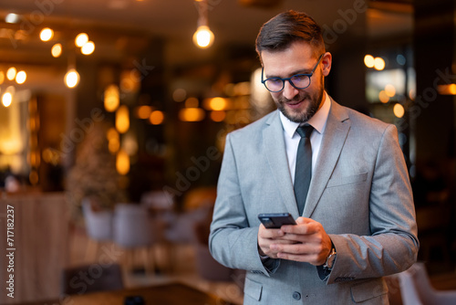 Young businessman using a smartphone in a co-working office. Modern businessman sending a text message to his business clients. Well dressed businessman standing alone in a modern workplace. photo