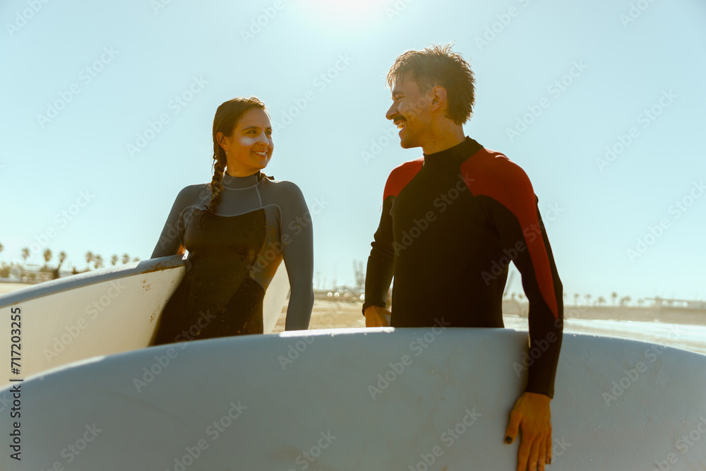 Couple of surfers in wetsuit with surfboards standing on the beach and looking each other with smile