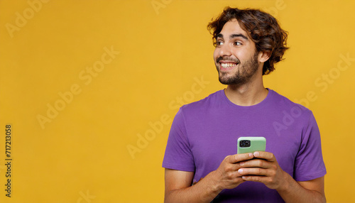oung man of African American ethnicity wear purple t-shirt casual clothes hold in hand use mobile cell phone look aside on area isolated on plain yellow background studio portrait. Lifestyle concept © Sam